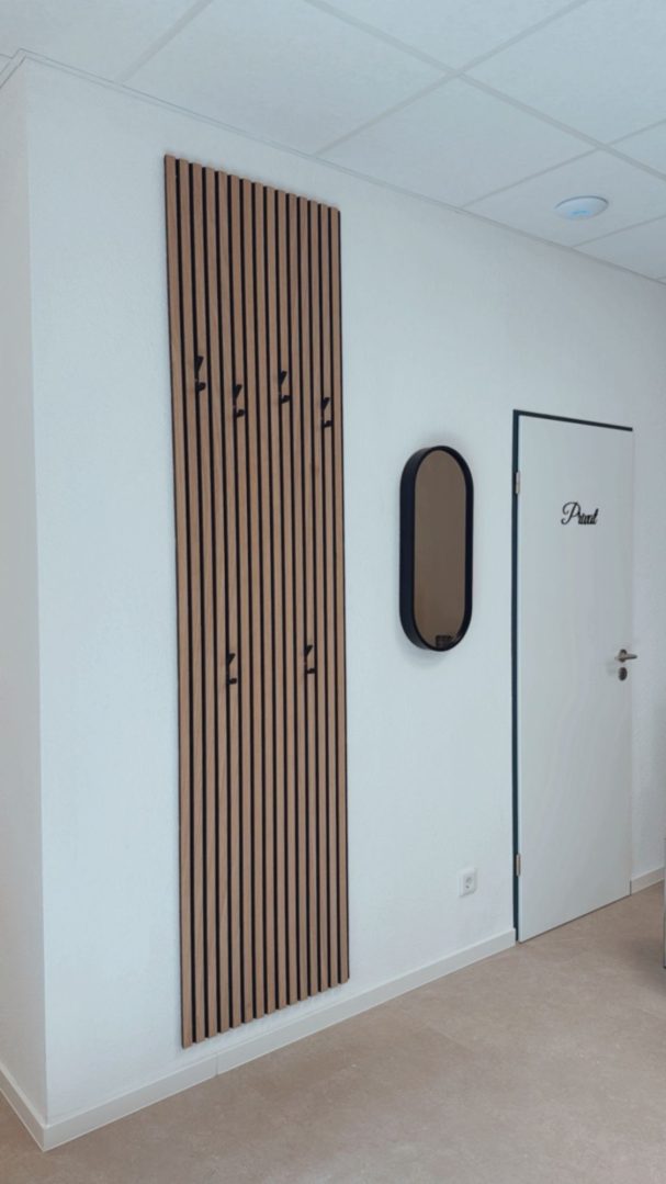 Holzregal mit Schlüssellöchern an einer Wand, daneben ein ovaler Spiegel und eine Tür.